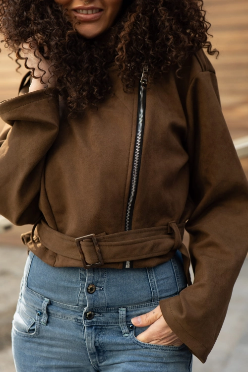 Detal brązowej kurtki ramoneski z ekozamszu: srebrny suwak i pasek z klamrą u dołu kurtki. Zbliżenie na strukturę materiału zamszowego.